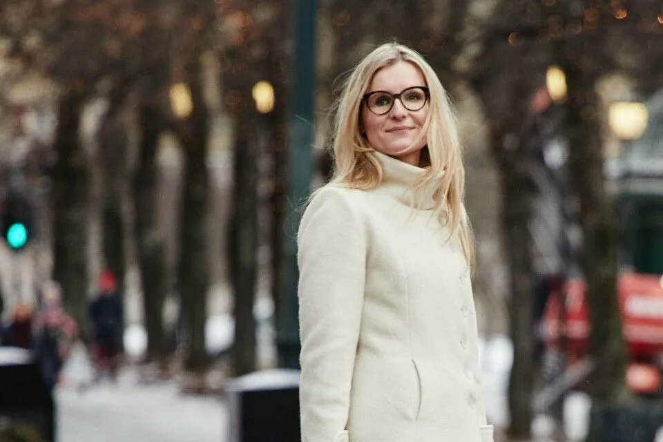 En blond person med briller og kremfarget frakk står utendørs i en trærom om vinteren.