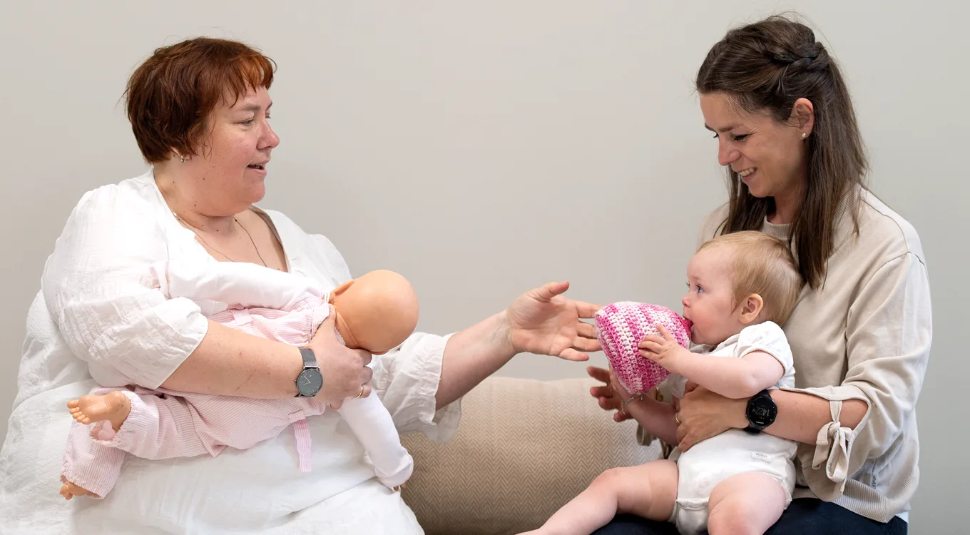 To personer i et lyst rom, den ene holder en dukke og den andre en baby, som samhandler med hverandre.