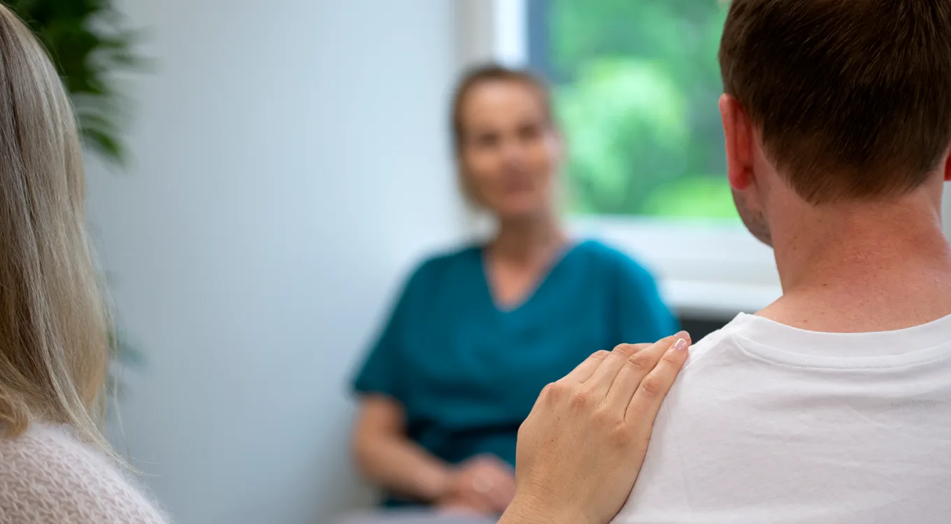 En person legger en trøstende hånd på en annens skulder i et uklart klinisk eller kontormiljø med en fagperson i nærheten.