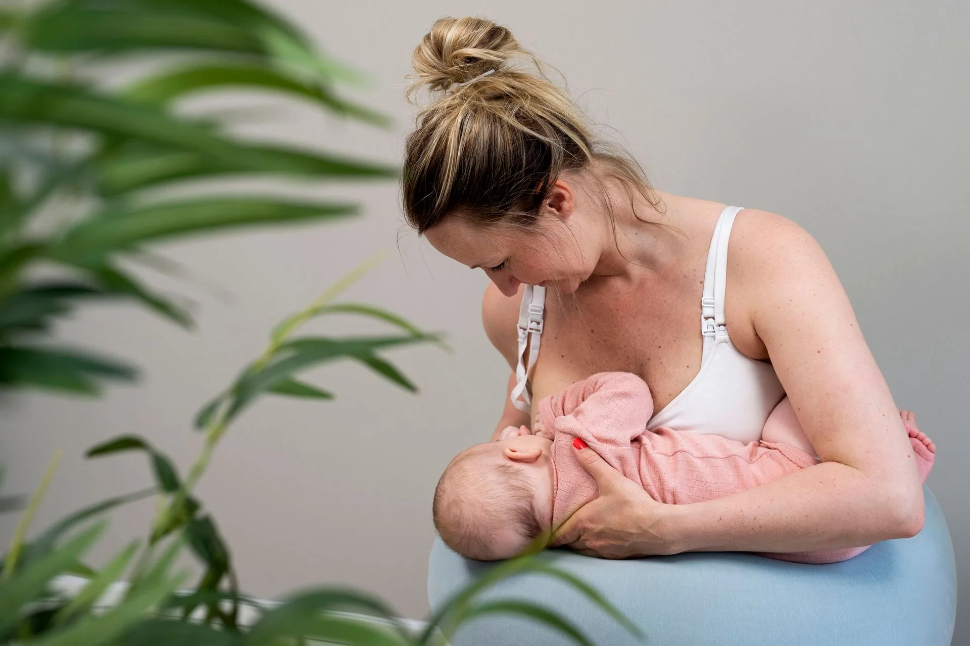 En person som sitter på en blå pute og ammer en baby, innrammet av grønne blader i forgrunnen.