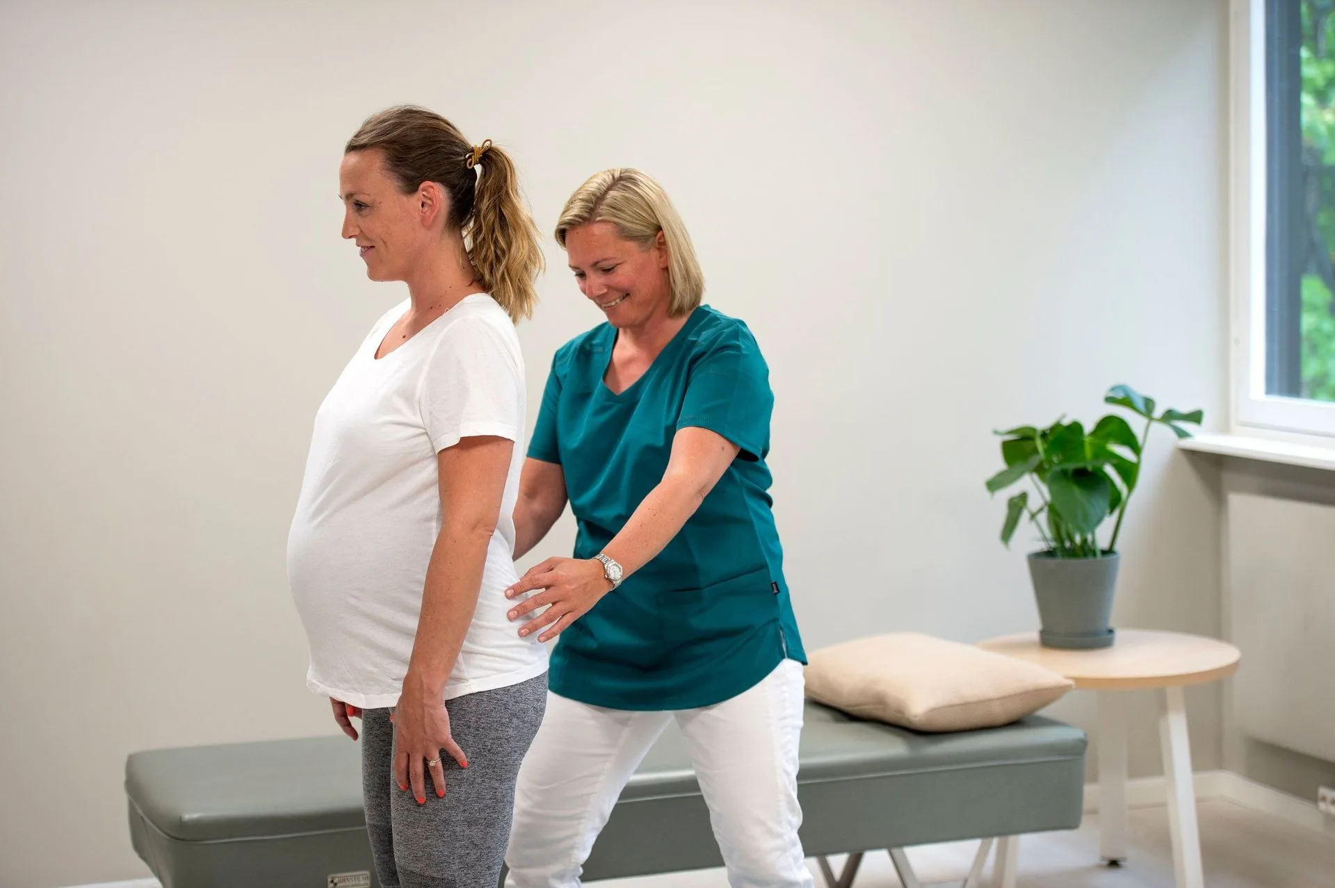 En fysioterapeut står bak en gravid pasient og veileder henne i holdningen hennes i et klinisk kontormiljø.