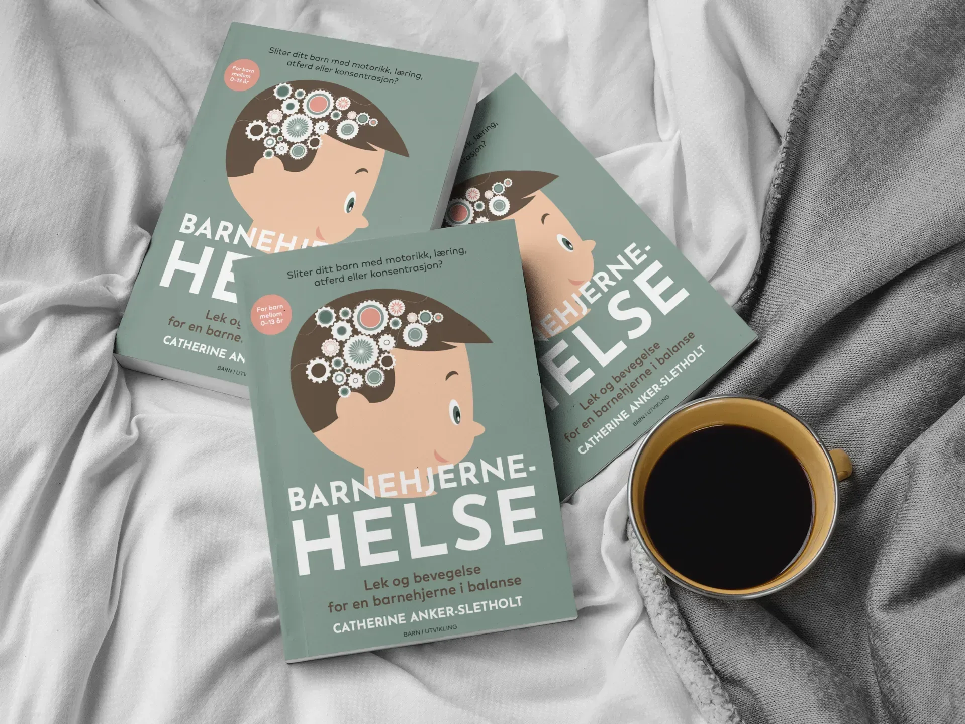 Tre eksemplarer av boken «Barnehjernehelse» arrangert på et hvitt ark ved siden av en kopp svart kaffe.