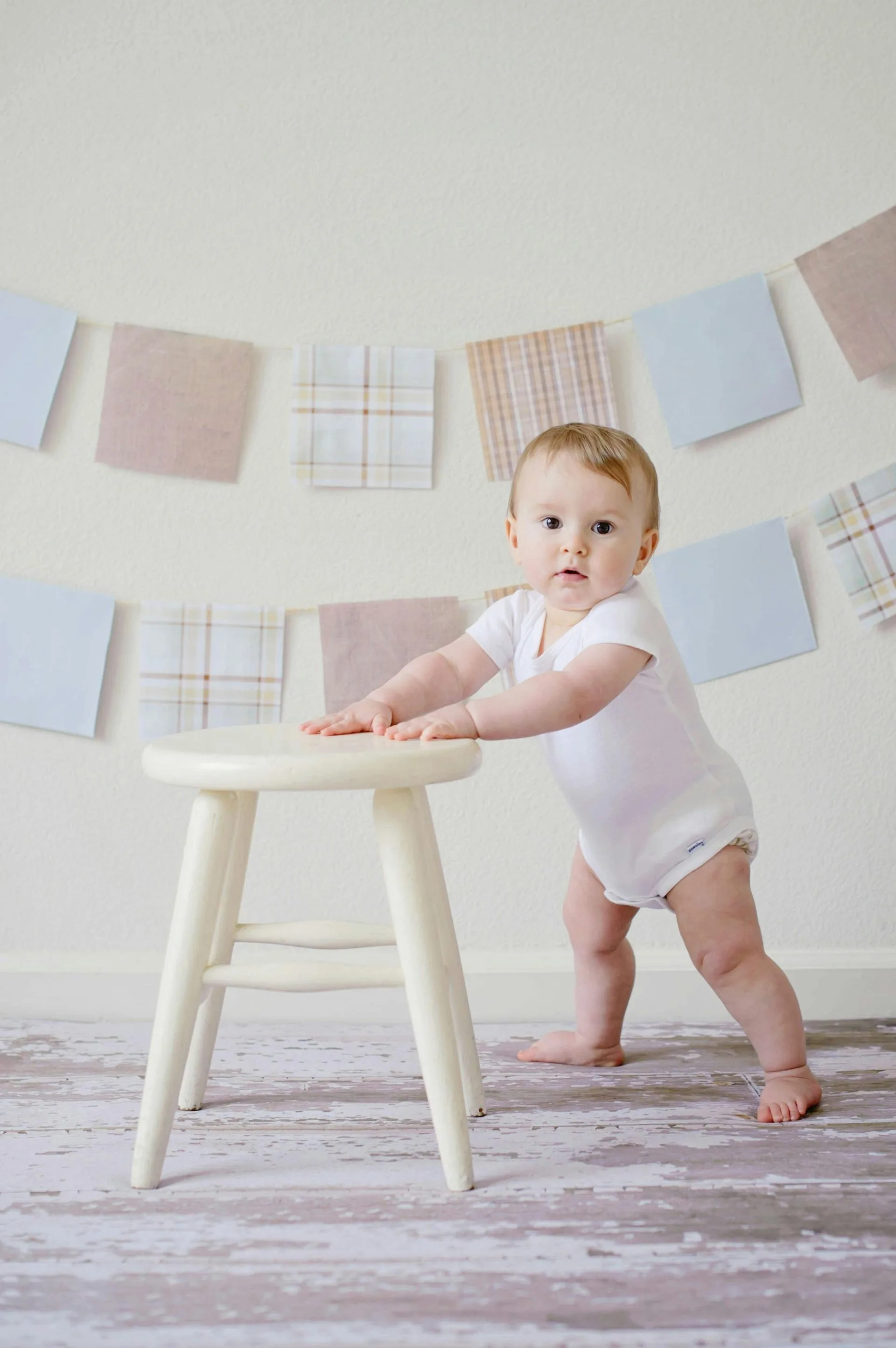 En baby som står og holder seg fast i en hvit trekrakk, med et dekorativt banner av stoffkvadrater i bakgrunnen.